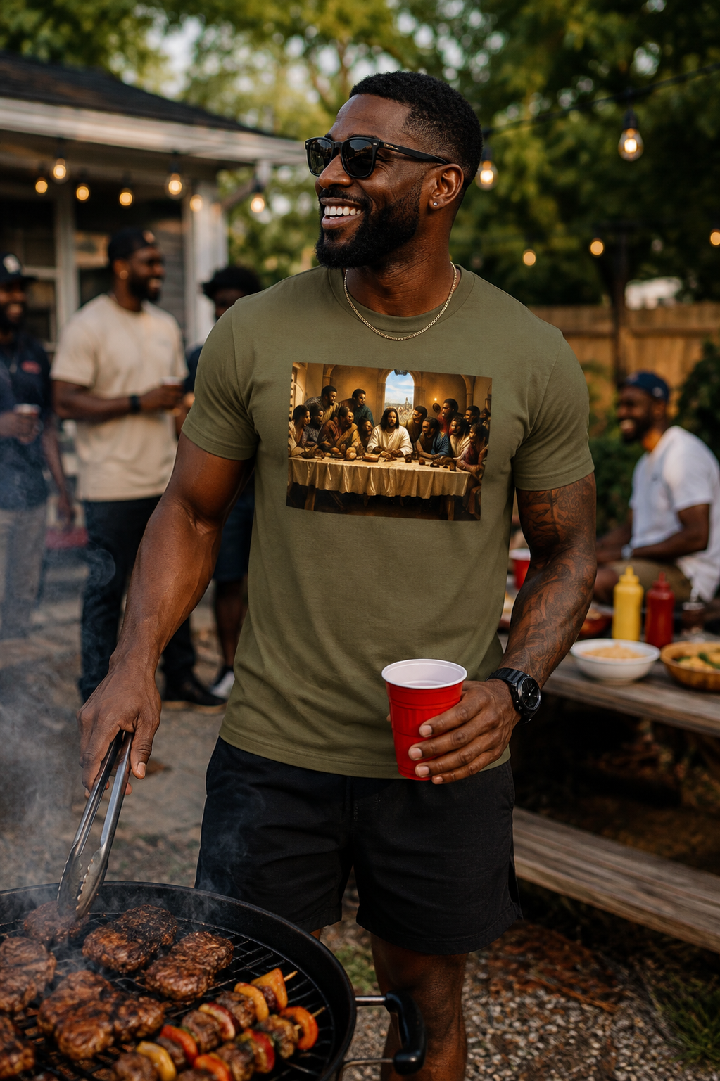 A man grilling at a backyard barbecue wearing an olive green t-shirt with a graphic print of Jesus and the disciples at the Last Supper, holding a red cup.